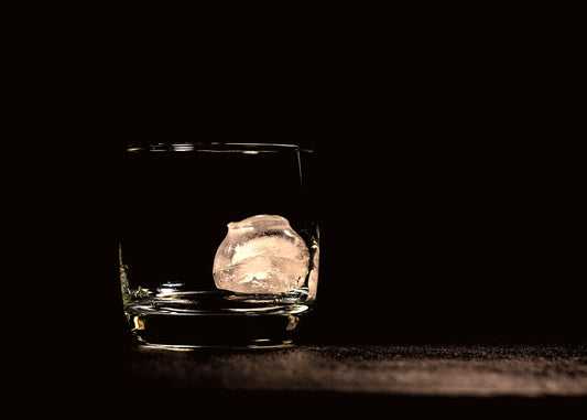Glass with one ice cube in it sitting on a brown surface with a black background 